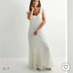 Vit blommig långklänning med volangärm - Romantisk vit långklänning med små blå och gröna blommor. Klänningen har volangärmar, v-ringning och knytning i ryggen. Tillverkad i ett lätt, transparent material som ger en luftig känsla. Perfekt för sommarens alla tillfällen.