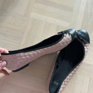 Beige quiltade ballerinaskor med svarta detaljer från H&M - Säljer ett par beige ballerinaskor från H&M med quiltat mönster och svarta tåpartier. Skorna har en liten rosett framtill och platt sula. Perfekta för en stilren och klassisk look.