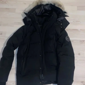 Svart dunjacka från Canada Goose med pälskant - Säljer en svart dunjacka från Canada Goose med huva och pälskant. Jackan har lång ärm, dragkedja och knappar framtill samt klassisk logotyp på ärmen. Perfekt för kalla dagar.