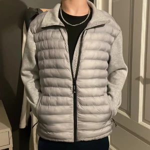 Ljusgrå pufferjacka med ärmar i sweatshirtmaterial - Säljer en ljusgrå pufferjacka med quiltad framsida och ärmar i mjukt sweatshirtmaterial. Jackan har dragkedja framtill och ståkrage. Perfekt för lager-på-lager och en stilren look.