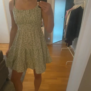 Grön blommig kortklänning med volang - Söt grön kortklänning med små vita blommor och volang nertill. Klänningen har smala axelband och en lätt utställd kjol, perfekt för varma dagar. Den har en feminin och somrig känsla med ett mjukt fall.
