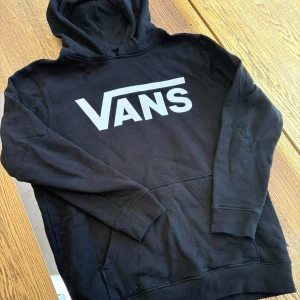 Svart hoodie från Vans - Säljer en svart hoodie från Vans med stor vit logga på bröstet. Klassisk modell med huva och magficka, perfekt för en avslappnad stil. Mjuk insida och ribbade muddar vid ärmslut och nederkant. ( storleken är junior large = XS)