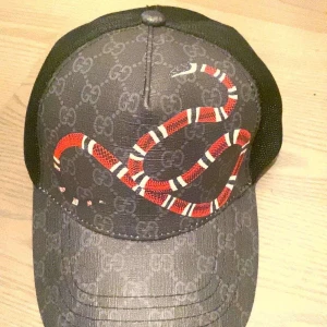 Svart keps med ormtryck från Gucci - Snygg svart keps från Gucci med diskret GG-mönster och en stor röd, vit och svart orm på framsidan. Kepsen har böjd skärm och mesh på sidorna för extra ventilation. Ej original men Perfekt för dig som vill sticka ut. 