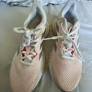 Adidas Solarglide vita och rosa löparskor - Säljer ett par Adidas Solarglide löparskor i vitt med ljusrosa detaljer och silvriga ränder på sidorna. Skorna har snörning, ventilerande mesh-ovandel och orange detaljer på sulan. Perfekta för dig som gillar snygga och bekväma träningsskor.