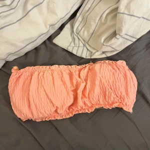 Ljusrosa bikinitopp - Säljer en ljusrosa bikini med smockad struktur och volangdetaljer upptill och nedtill. bikinin är ärmlös och har en mjuk, stretchig passform som sitter bekvämt runt kroppen. Perfekt för varma dagar eller sommarens festivaler! 