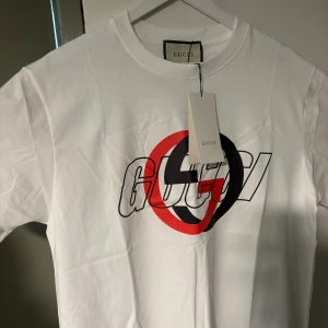 Vit t-shirt  - Säljer en vit t-shirt  med ett stort grafiskt tryck i rött och svart framtill. Klassisk rund halsringning och korta ärmar. Perfekt för dig som gillar stilrena plagg med en twist. Perfekt till sommaren ☀️ 🏝️ Varan är i nyskick & etikett medföljer samt inplastad i  påse av märket!  STRL M men den är lite baggy i passformen så den passar ÄVEN de som pendlar mellan strl M & L! Varan är i nyskick så respektera gärna priset.