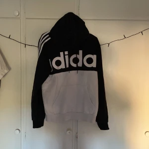 Svart och vit hoodie från Adidas - Säljer en svart och vit hoodie från Adidas med stor logga framtill och klassiska tre ränder på axlar och ärmar. Tröjan har huva och magficka, samt ribbade muddar vid ärmslut och nederkant. Perfekt för en sportig look!