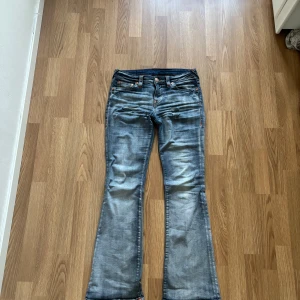 Blå bootcut jeans från True Religion - Säljer ett par klassiska blå jeans från True Religion med bootcut-passform. Jeansen har tydliga kontrastsömmar, stora bakfickor med signaturbroderi och detaljerade nitar. Perfekta för dig som gillar en tidlös denimlook.