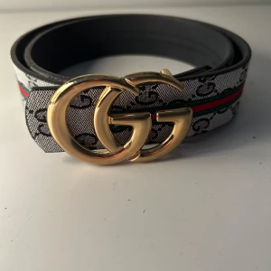 Bälte med GG-spänne - Snyggt bälte med stort guldfärgat GG-spänne. Bältet har ett mönstrat band i grått och svart med röda och gröna detaljer längs kanten. Tillverkat i skinn med klassisk design
