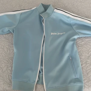 Ljusblå track jacket från Palm Angels - Säljer en ljusblå track jacket från Palm Angels med vit logga på bröstet. Jackan har vita och svarta ränder längs ärmarna, dragkedja framtill och fickor med dragkedja. Tillverkad i polyester för en sportig känsla.