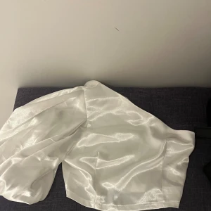 Vit glansig kortkjol med dragkedja - Säljer en vit glansig topp i satinliknande material. Toppen har en dragkedja vid sidan och en stilren, enkel design utan mönster. Nästan oanvänd!