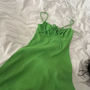 Grön klänning med smala axelband - Säljer en snygg grön klänning med smala axelband och knytning framtill. Klänningen har en enkel och stilren design med lätt rynkad struktur i tyget. Perfekt för varma dagar!