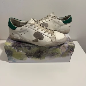 Vita sneakers från The Gallerist med gröna detaljer - Säljer ett par vita sneakers från The Gallerist med gröna hälpartier och ett stort svart klövertryck på sidan. Skorna har klassisk snörning och platt sula, perfekt för en stilren och cool look. Priset är inte hugget i sten!