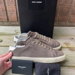 Saint Laurent skor - Säljer ett par snygga beige sneakers från Saint Laurent i mjuk mocka med vita detaljer vid hälen och vit platt sula. Klassisk design med snörning och diskret logga på sulan. Perfekta för en stilren vardagslook.