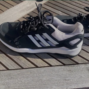 Svarta Adidas sneakers med vita detaljer - Säljer ett par svarta Adidas sneakers med klassiska vita ränder på sidorna och vit sula. Skorna har snörning och en sportig design med ventilerande meshfoder. Perfekta för vardagsbruk eller träning.