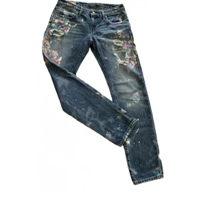Målade boyfriend jeans från Polo Ralph Lauren - Unika blå boyfriend jeans från Polo Ralph Lauren med färgstänk och slitningar. Jeansen har raka ben, låg midja och detaljerade hål samt färgfläckar för en kreativ look. Perfekta för dig som vill sticka ut med din stil. Använda 3 gånger