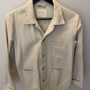 Beige vårjacka från A Day's March - Stilren beige vårjacka från A Day's March med klassisk krage och tre utanpåfickor framtill. Jackan har knäppning med knappar. 