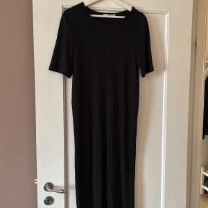 Svart ribbad klänning från Ellos - Svart ribbad långklänning från Ellos i storlek 42/44. Klänningen har korta ärmar, rund halsringning och är i en rak, enkel modell som går ner till vaden. Perfekt för en stilren och bekväm look.