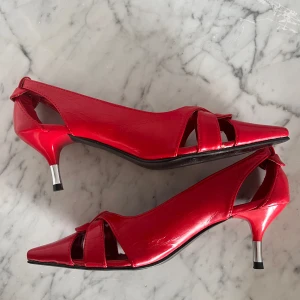 Röda klackskor med öppen tå och spetsig tå - Säljer ett par röda klackskor med öppen tå och spetsig tå. Skorna har snygga utskärningar på sidorna och en smal klack med silverdetalj. Tillverkade i glansigt syntetmaterial och passar perfekt för dig som vill sticka ut.