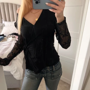 Svart omlottopp med spetsärmar - Snygg svart omlottopp med transparenta spetsärmar och knyt i sidan. Toppen har v-ringning och är perfekt för att lyfta vilken outfit som helst. Passar dig som gillar en lite mer elegant och trendig stil.