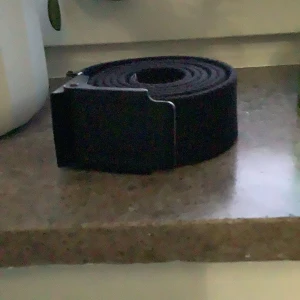 Svart tygbälte med metallspänne - Säljer ett svart bälte i tyg med ett stilrent metallspänne. Bältet är smalt och passar perfekt till jeans eller chinos. Enkel design utan mönster eller logga, vilket gör det lätt att matcha med olika outfits.