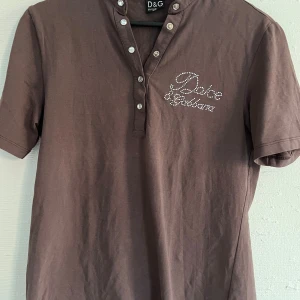Brun t-shirt från Dolce & Gabbana - Brun t-shirt från Dolce & Gabbana med korta ärmar och dekorativa knappar vid halsen. Framsidan har logga i strass och baksidan har ett stort tryck med krona och logga i silver. Perfekt för dig som gillar statement-plagg.