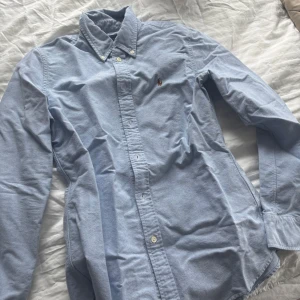 Ljusblå skjorta från Polo Ralph Lauren - Klassisk ljusblå skjorta från Polo Ralph Lauren med button-down krage och broderad logga på bröstet. Skjortan har långa ärmar och vita knappar framtill. Perfekt till en stilren och avslappnad look.