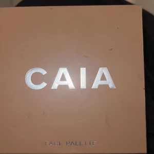 CAIA Face Palette - En sminkpalett från CAIA med fyra olika puder: contour i en kall brun nyans, blush i stark rosa, bronzer i varm brun och highlighter i skimrande persikoton. Perfekt för att skapa en komplett bas med både lyster och färg.