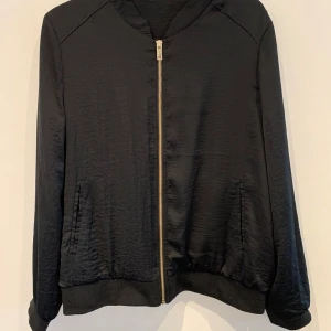 Svart bomberjacka med dragkedja - Säljer en stilren svart bomberjacka med silkeslen yta och dragkedja framtill. Jackan har långa ärmar, ribbade muddar och två fickor på sidorna. Perfekt för dig som vill ha en enkel men snygg jacka till vardags.