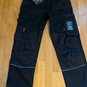 Svarta arbetsbyxor från Jobman - Svarta arbetsbyxor från Jobman med flera praktiska fickor, bland annat benficka med plastfönster för ID-kort. Byxorna har förstärkta knän och reflexdetaljer vid bensluten. Perfekta för dig som behöver tåliga byxor med smarta funktioner. Helt nya 