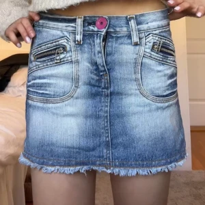 Cool jeanskjol - Säljer en kort blå jeanskjol med fransad nederkant och två dragkedjefickor framtill. Kjolen har en rosa knapp i midjan och markerade sömmar som ger en cool look. Perfekt för dig som gillar denim och vill ha något trendigt i garderoben.