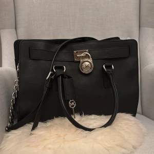Svart handväska från Michael Kors - Snygg svart handväska från Michael Kors med silverfärgade metalldetaljer och kedja på sidan. Väskan har både handtag och axelrem samt MK-logga framtill. Perfekt för dig som vill ha en stilren och klassisk accessoar.
