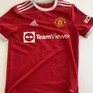 Manchester United matchtröja Elanga 36 - Röd Manchester United fotbollströja med Elanga och nummer 36 på ryggen. Adidas-logga och TeamViewer sponsortryck på framsidan. Klassiska vita detaljer vid krage och ärmslut samt tre ränder på axlarna.