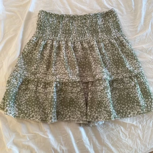 Mini kjol - Supersöt grön kjol med vitt blommönster och volanger. Kjolen har smock i midjan för en bekväm passform och är tillverkad i ett lätt, luftigt material. Perfekt för dig som gillar romantisk och trendig stil. Midjebandet går att vika ner och göra kjolen ännu mera lågmidjat, midjebandet är även stretchigt. Skriv via funderingar eller mera bilder💕