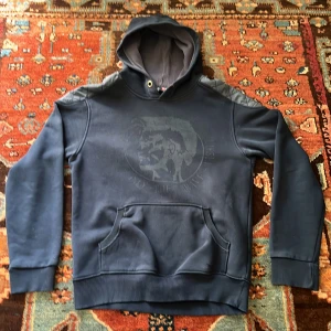 Mörkblå Diesel hoodie  - Mörkblå Diesel hoodie med stort tryck på bröstet. Tröjan har quiltade detaljer på axlarna, känguruficka och huva med snörhål. Perfekt för en relaxed fit