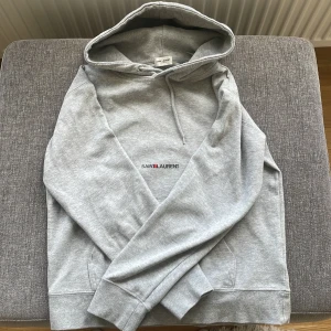 Grå hoodie från Saint Laurent - Säljer en stilren grå hoodie från Saint Laurent med diskret logga broderad på bröstet. Tröjan har huva med dragsko och en klassisk känguruficka framtill. Perfekt för en avslappnad och trendig look.