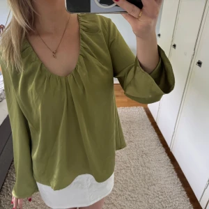 Olivgrön blus från NA-KD - Säljer en olivgrön blus från NA-KD med lång ärm och smickrande passform. Blusen har rundad halsringning med rynkade detaljer och är tillverkad i ett mjukt, lätt material. Perfekt för en stilren och avslappnad look.