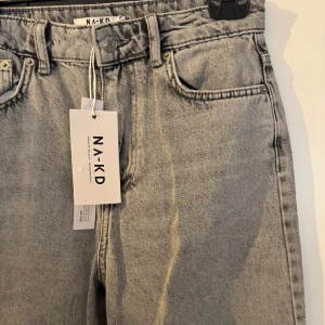 Gråa jeansbyxor från NA-KD - Säljer ett par gråa jeansbyxor från NA-KD i klassisk femficksmodell. Byxorna har hög midja och raka, vida ben.