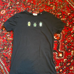 Svart t-shirt med skalbagge-tryck från Lindbergh - Svart t-shirt från Lindbergh med tryck av tre färgglada skalbaggar på bröstet. Klassisk rund halsringning och korta ärmar. 