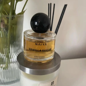 NAIYA Vanilla Sugar parfym - Parfymflaska med doften Vanilla Sugar från NAIYA. Flaskan är rund och genomskinlig med svart klotformad kork och bär en varm, gyllene vätska. Snygg och stilren design som passar perfekt på sminkbordet.