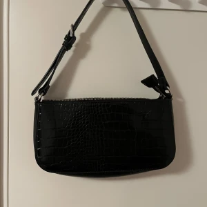 Svart axelväska i krokopräglat skinn - Snygg svart axelväska med krokopräglat mönster och justerbart axelband. Väskan har dragkedja upptill och ett rymligt fack inuti. Perfekt storlek för det viktigaste och ger en cool vibe till din outfit.Katt finns i hemmet 
