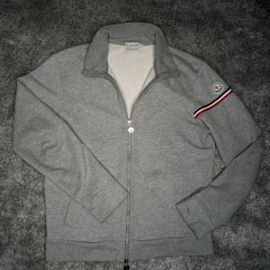 Moncler ziptröjä - Snygg grå ziptröja från Moncler med klassisk ståkrage och hel dragkedja. Tröjan har lång ärm, broderad Moncler-logga på ena ärmen och en röd, vit och marinblå rand som detalj. Tillverkad i mjuk bomull för en skön känsla.