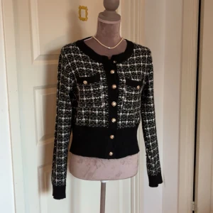 Svartvit rutig kofta med guldknappar - Snygg svartvit rutig kofta med guldiga knappar framtill och på fickorna. Koftan har lång ärm, ribbade muddar och en croppad passform. Perfekt för dig som gillar en chic och trendig look. Materialet känns mjukt och stickat.