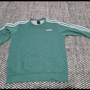 Grön Adidas sweatshirt med vita ränder - Säljer en grön elastisk sweatshirt från Adidas i storlek M, fits också på L män. Tröjan har klassiska vita ränder längs ärmarna och Adidas-logga på bröstet. Mjuk och skön med rund hals och långa ärmar, perfekt för en sportig look.