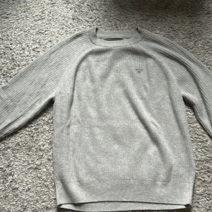 Grå stickad tröja från GANT - Säljer en stilren grå stickad tröja från GANT med rund hals och diskret logga broderad på bröstet. Tröjan har ribbade muddar och är långärmad, perfekt för lager-på-lager under kyliga dagar.