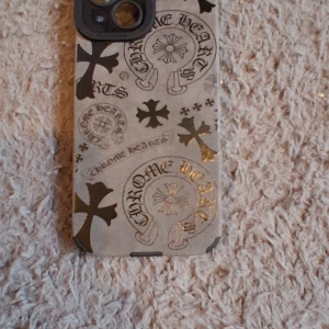 Chrome Hearts iPhone 15 skal - Snyggt iPhone 15 skal från Chrome Hearts med grå bas och svarta samt guldiga kors och loggor över hela baksidan. Skalets design är riktigt edgy och sticker ut med sina detaljerade tryck och metallic-effekter. Perfekt för dig som vill ha en unik vibe på mobilen.