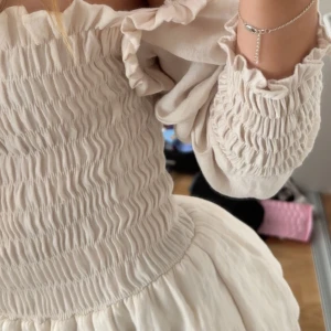 Ljus offshoulder kortklänning med volang - Supersöt ljus kortklänning med offshoulder och smockad överdel. Klänningen har långa puffärmar, volangdetaljer och en flowy kjol. Materialet känns svalt och luftigt.!!!! har klippt av den sista volangen på kjolen inget som syns när man har på den.!!!! Från Jubil Studio