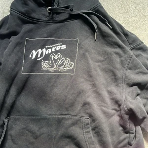 Svart Mares hoodie med turnétryck - Svart hoodie från Mares med vit tryckt logga och svanmotiv på bröstet samt turnélistan 'Svanesång Tour 2023' på ryggen. Klassisk känguruficka framtill, huva med snörning och mjukt bomullsmaterial. 