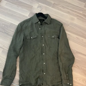 Olivgrön linneskjorta från Massimo Dutti - Snygg olivgrön skjorta från Massimo Dutti i linne. Klassisk modell med två bröstfickor med lock och vita knappar framtill. Skjortan har lång ärm och avslappnad passform, perfekt för en stilren och chill look. Även bra som en overshirt nu mot hösten. 