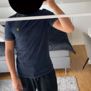 Mörkblå t-shirt från Lyle & Scott - Mörkblå t-shirt från Lyle & Scott med gul logga på bröstet. T-shirten har korta ärmar och ribbade muddar med märkets namn broderat i ljusblått. Skön passform och tillverkad i mjuk bomull.
